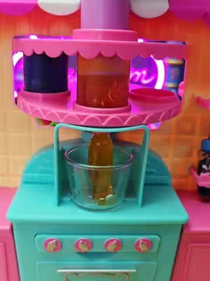 The image shows a colorful, toy-like contraption with a teal countertop, pink accents, and a multi-tiered structure. In the center is a clear cup positioned under a container that appears to be dispensing a golden liquid. The background features a lighted, blurred environment.