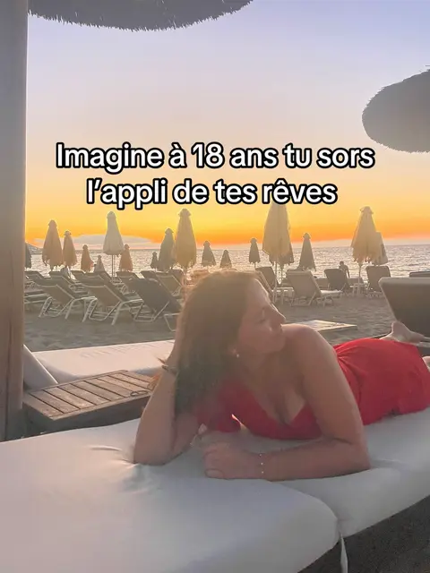 A woman wearing a red dress is lying on a white lounge chair on a beach. Behind her is a sunset over the ocean, with beach umbrellas creating a backdrop.