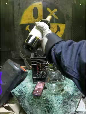 The image shows a scene where a person, wearing safety gloves and a dark jacket, is holding a bottle of champagne. The bottle is positioned as if the person is about to smash it in the frame, with a shattered glass surface in the foreground and a background with a logo design on a black surface. There are also some objects in the scene, including a black speaker, a phone with a screen illuminated, and a clear glass container.