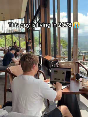 The cover photo shows a man sitting at a table indoors, working on a laptop, with a sophisticated setting in the background. The man is seen from the back, focusing on the laptop screen. Behind him, large windows offer a view of a bright, sunny environment with mountains and green trees. There are other people visible in the background, some seated and some standing, enjoying the ambiance.
