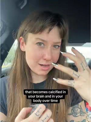 A woman with long brown hair and a side bang is seated in a car, looking towards the camera. She has fair skin, is gazing with a subtle smile, and is adorned with a nose piercing and long, silver and black nail polish. Her left hand is raised, fingers extended, with a tattoo on two fingers. She has a dark-colored top and a tattoo on her upper arm which is visible. A text overlay appears in a black rectangular box at the bottom.