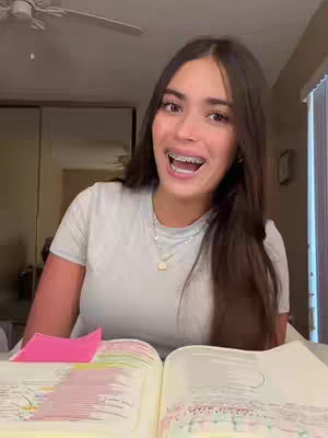 A woman is sitting at a table with an open book, smiling and looking directly at the camera. She has long dark hair and is wearing braces. The background appears to be a home setting with neutral colors.