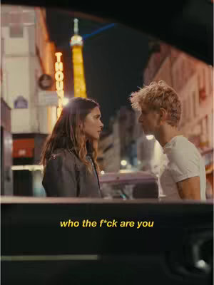 Two people are facing each other in an urban setting at night. A woman with long hair is on the left, and a man with blonde hair is on the right. The foreground is dark, possibly the interior of a car, and in the background, there is a tower with a yellow neon sign and buildings. The sky is dark. Text overlayed at the bottom reads 'who the f*ck are you'.