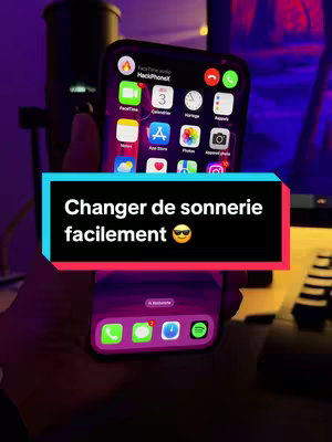 A person's hand holding an iPhone displaying the home screen. A text overlay is in a black and red box positioned on the phone screen. The background is dimly lit with a microphone.