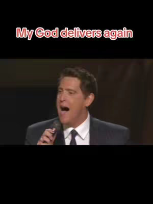 The image shows a man in a dark suit, holding a microphone and speaking with an open mouth, likely giving a speech. Above him, in a contrasting color, is the text overlay "My God delivers again."