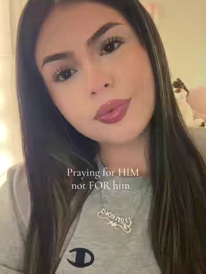 A selfie featuring a young woman with long, dark hair, posed slightly angled towards the camera. She has full, glossy lips and is wearing a silver pendant necklace. The text overlay reads 'Praying for HIM not FOR him.' in a casual font. The background appears to be a lightly blurred indoor setting, with a soft, warm color palette.