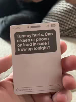 A hand holding a card designed to look like a phone screen. The screen shows a text message and a phone interface, revealing a casual conversation. The background is blurred, hinting at a domestic setting.