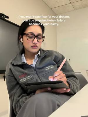 A woman wearing glasses, a gray zip-up jacket, and a light striped shirt is focused on a tablet she is holding in her lap. She has earbuds in her ears and is holding a pink stylus. The background is a blurred office setting. Text overlay: 'If you won't sacrifice for your dreams, don't be surprised when failure becomes your reality.'