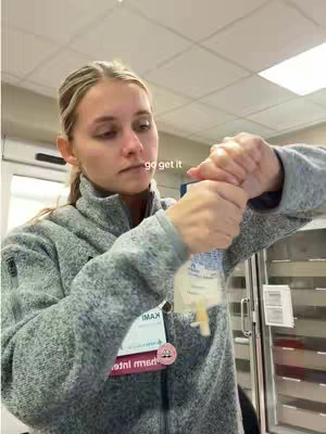 A woman in a grey zip-up jacket is holding a medical bag or device. She is in a medical setting, possibly a clinic or hospital, with the background including a door and some medical equipment.