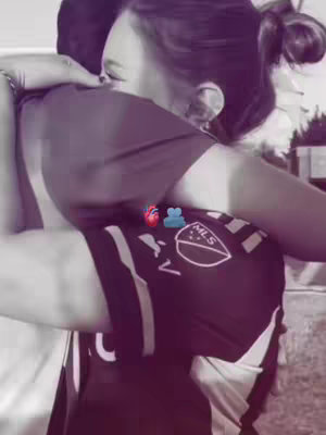 A grayscale image shows two people hugging. The person on the left is obscured by the other person's back. The person on the right is a woman with her hair in a bun, her face partially visible, and her eyes closed. She is wearing a dark shirt, and the other person is also wearing dark clothes. A jersey with an MLS logo is visible. The image is soft and emotional, with minimal background detail.