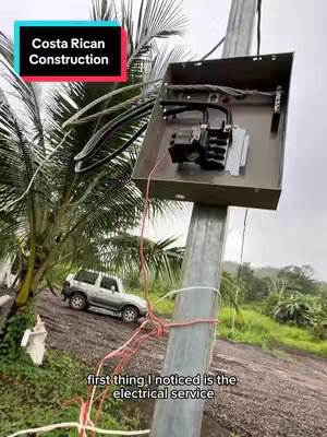 The cover image features an electrical box mounted on a metal pole, with wires and other components visible. Text overlays indicate that the video is about Costa Rican construction. In the background, a white SUV is parked on a dirt road with lush green foliage.