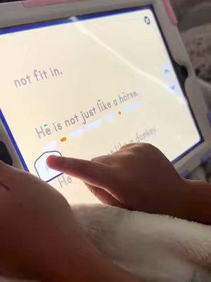 A close-up shot of a child's hands interacting with a tablet screen displaying text from a reading app. The tablet is held at an angle, with the child's fingers pointing toward the screen. The background is out of focus and appears to be a bed or soft surface.