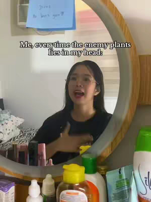 A young woman in a black shirt and glasses stands in front of a round mirror. The mirror reflects a room with various items on a shelf below, including cosmetics and toiletries. A blue sticky note with a handwritten message is stuck on the mirror. Above, text overlaid on the image reads, "Me, everytime the enemy plants lies in my head:".