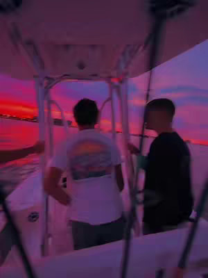 A low-angle shot from inside a boat, looking out at a vibrant sunset. Two young men are in the frame, with the focus slightly blurred. One is standing near the boat's control area, and the other is facing toward the setting sun. The sky is painted with intense hues of purple, pink, and orange. The boat's structure is visible, with support beams and a covered area overhead.