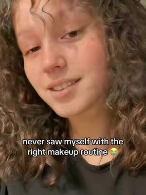 A selfie-style shot features a person centered within the frame. She has curly hair framing her face, and her expression is a slight smile. The background is blurred, suggesting it's an indoor setting.