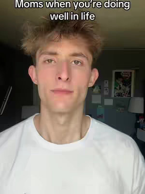 A medium shot captures a young man with light skin and tousled, light brown hair in the foreground, centered in the frame. He wears a simple white t-shirt. Framing the shot, the background is dimly lit, suggesting a home interior. The image has a casual, personal feel thanks to the close-up style and simple lighting.