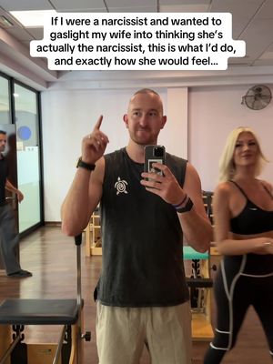 A man in a tank top takes a selfie, pointing upwards with his right index finger, while a woman performs an exercise nearby. The background is a light-colored room with exercise equipment. Text is added on top of the image.