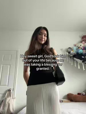 A woman is standing indoors, dressed in a black top and a long pleated skirt, facing the camera. The background shows a door, a wall, and a bed with scattered items, including stuffed animals.