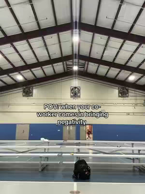 The image shows the interior of a large indoor sports facility with rows of metal benches in the foreground and a basketball court in the background. The text overlay reads 'POV: when your co-worker comes in bringing negativity'.