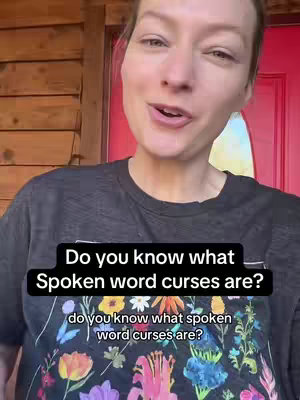 A woman is filmed from the chest up, talking to the camera with a serious expression. She is standing inside a wooden door frame with dark hair, wearing a dark t-shirt with a flower design. Behind her is a door painted red, with a portion of a light blue wall visible.