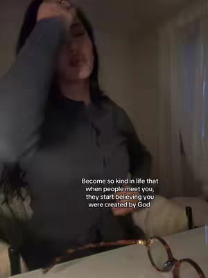 The image captures a woman from the chest up, indoors and slightly blurred. She has black hair and is wearing a dark gray top with a hint of her face visible. Text overlays on the images with the phrase 'Become so kind in life that when people meet you, they start believing you were created by God.' Additionally, glasses are visible at the bottom. The lighting is dim, creating a somewhat intimate atmosphere.