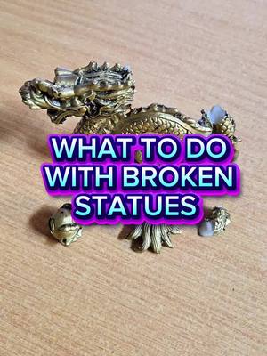 The cover image shows a light-brown wooden surface. On it, there's an ornate, golden statue of a dragon with its head raised. Superimposed over the statue is text in a vivid combination of pink, purple, and blue with white outlines that reads: 'WHAT TO DO WITH BROKEN STATUES.' The dragon statue has been apparently broken and has a couple of missing pieces, spread below. The composition uses a close-up perspective with the statue positioned centrally.