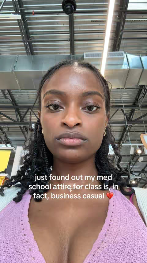 A selfie showing a young woman from the chest up, facing the camera. She has dark skin, dark curly hair styled in braids, and is wearing a pink top. The background is a generic indoor setting with industrial-style ceilings and lighting. Text overlay at the bottom of the image.