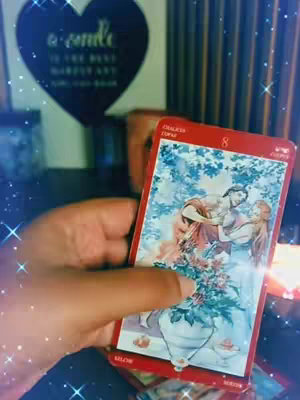 A hand holding a tarot card in front of a blurred background. The card is red with an illustration of two figures embracing and an overflowing vase, blue and white elements. The background includes a black heart-shaped sign with script, and vertical brown wood tones.