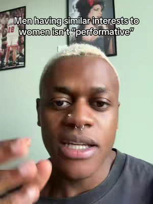A close-up shot of a Black man with short, blonde-tipped hair and a nose ring, looking directly at the camera. He appears to be speaking, with his mouth open slightly and his hand and arm are visible in the bottom left. Behind him, the wall has a light green hue, and two framed pictures hang on the wall. The one on the left appears to be of a man in basketball gear, and the one on the right is of a woman.