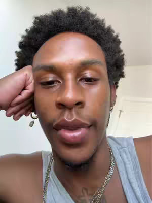 A close-up shot of a person with dark skin and curly black hair. The person is looking down and slightly to the left, resting their right hand on their head. They are wearing a gray tank top and a silver chain necklace. Dangling earrings are visible on their left ear. The background is a plain white wall.