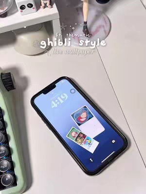 The cover image showcases a phone on a white desk with a Ghibli-themed live wallpaper. The phone, encased in a black case, displays a custom lock screen featuring a Ghibli character illustration and widgets. Above the phone, other Ghibli figurines and decorative elements, like a tiny typewriter, accentuate the aesthetic. The background is light-colored, and the layout features the phone prominently in the center.