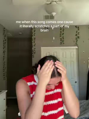 A person with dark hair wearing a red and white vertically striped tank top stands in a bedroom, seemingly reacting to something. The person has their hands on their head and appears to be looking down with a slight frown. Behind the person is a white door, a ceiling fan, and vines hanging from the ceiling. The shot suggests a personal, relatable moment.