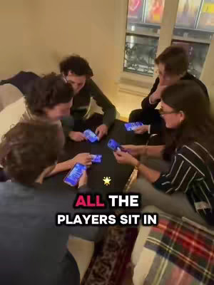The cover image shows a group of five young people gathered around a low table, likely in a living room setting. They appear to be engaged in a game, each holding a smartphone with a glowing blue screen. The color palette is warm, with soft lighting illuminating the scene. A text overlay at the bottom states, "ALL THE PLAYERS SIT IN."