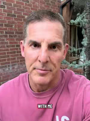 A man in a pink shirt is the focus of a selfie-style video. He stands in front of a brick wall and beside some leafy green bushes or trees. At the bottom of the image, there is a text overlay that says "WITH ME" in white font with a black border.