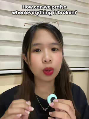 A woman with a bright complexion and red lipstick is the focus of the image, her gaze directed towards the viewer while her lips appear open. The background includes light-colored, horizontally striped blinds, with a pair of glasses perched above her head. Her dark hair frames her face, and she's wearing a black top with a necklace featuring a pendant.