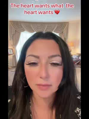 A selfie-style video cover featuring a woman with long, dark hair and heavy eye makeup. The background includes a beige curtain and hints of an interior space. Text overlayed across the top reads "The heart wants what the heart wants ❤️".