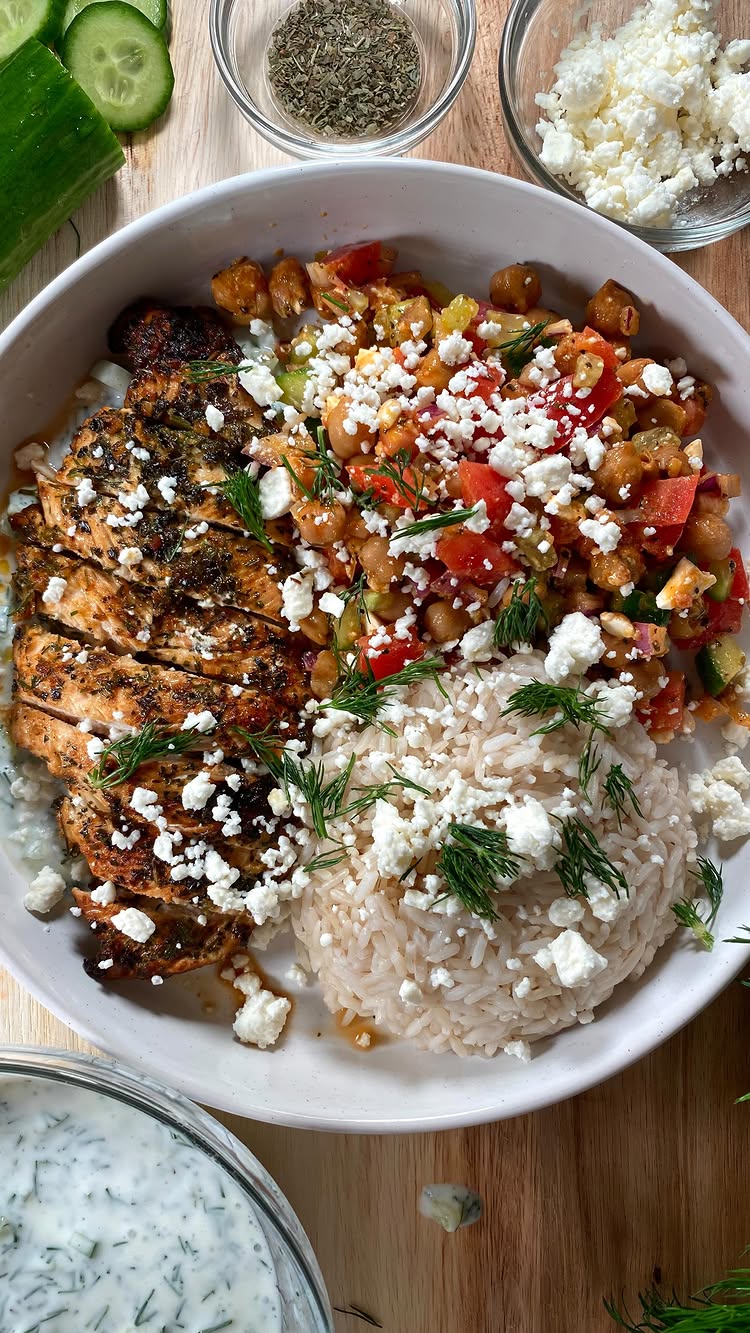 An overhead shot of a Mediterranean chicken plate. The plate is filled with grilled chicken bathed in herbs, rice, chickpeas, tomatoes, cucumbers, and feta cheese, beautifully arranged and presented on a light wooden surface with small bowls of herbs and cheese.