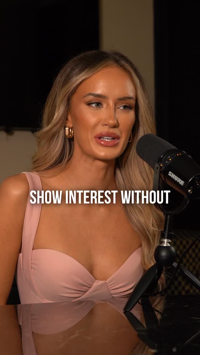 A medium shot features a woman speaking, positioned slightly off-center with a microphone to her right. She is wearing a pink dress with a sweetheart neckline. The text overlay reads 'SHOW INTEREST WITHOUT'. The background is dark.