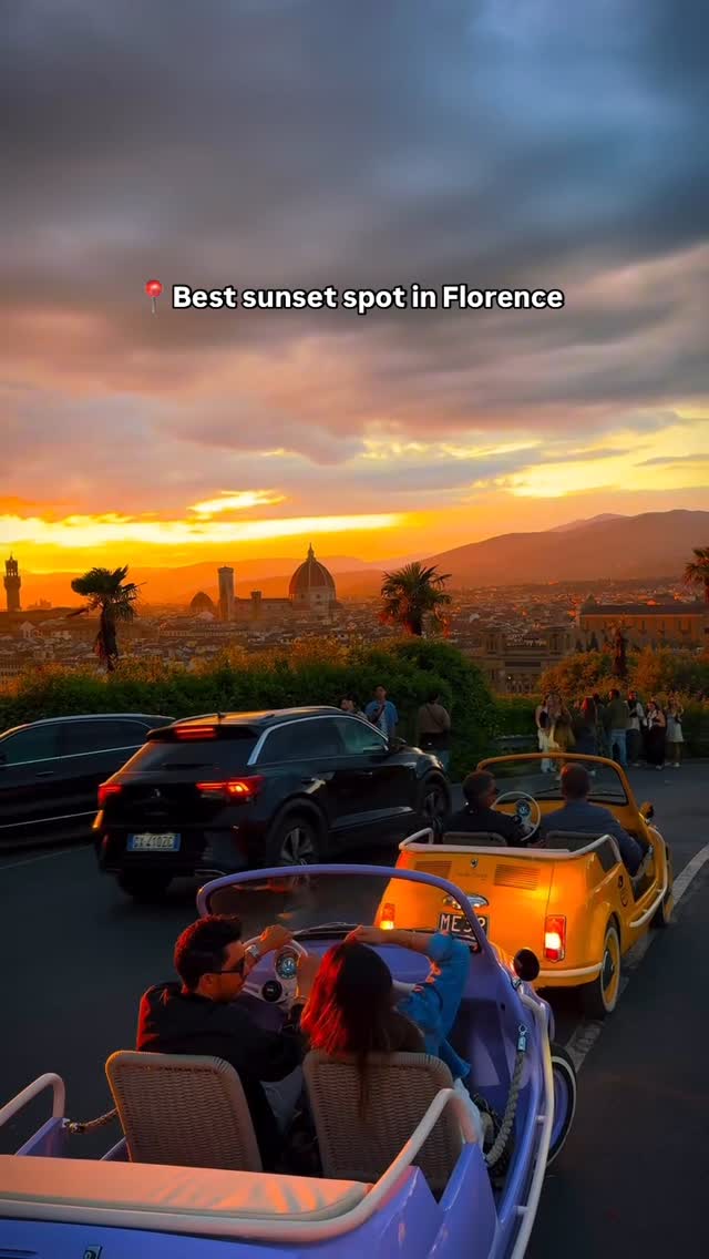 The image captures a stunning sunset over Florence, Italy. The warm, golden light bathes the cityscape, highlighting iconic landmarks like the Duomo. In the foreground, colorful vintage cars, including a yellow Fiat 500 and a purple open-top car, are filled with people, adding movement and a sense of joy to the scene. Silhouetted dark cars add depth, and a group of people stand and watch the sunset creating depth. The sky is a blend of oranges, pinks, and purples, suggesting a vibrant, late-afternoon hour.