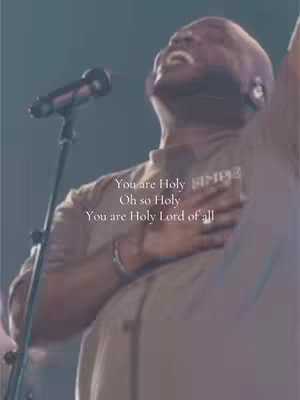 A man with dark skin sings passionately into a microphone, his face tilted upwards with his eyes closed. Text overlays the image, with the words 'You are Holy' in white with a soft background.