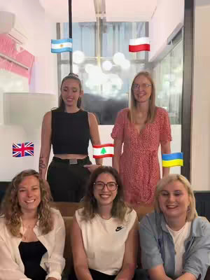 Six young women are gathered in a modern, well-lit space. Some are seated on a tan bench, while others stand behind them. Each woman holds a small flag of a different country above their heads. The flags include the United Kingdom, Ukraine, Poland, Argentina, Lebanon and Ukraine.
