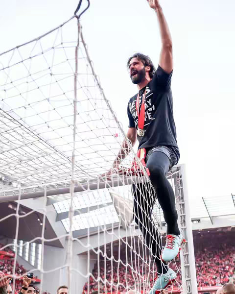 A wide shot captures a soccer goalkeeper celebrating a victory. He is positioned on a net structure, raising his arm in triumph. He wears a black shirt revealing a message, a gold medal, with fans visible in the background, suggesting a stadium setting.