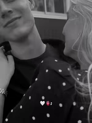 A black and white image featuring a man and a woman. The man, on the left, has his face turned towards the woman, with a slight smile. The woman is on the right, with her face near the man's, partially out of frame. The man is wearing a dark shirt, and the woman is wearing a polka dot top. Small heart and cherry emojis overlay the image.