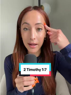 A woman with auburn hair and orange nails is holding a microphone and touching her eyebrow with her finger, while facing the camera, smiling slightly. A text overlay on a colored background that reads "2 Timothy 1:7" is below the microphone.