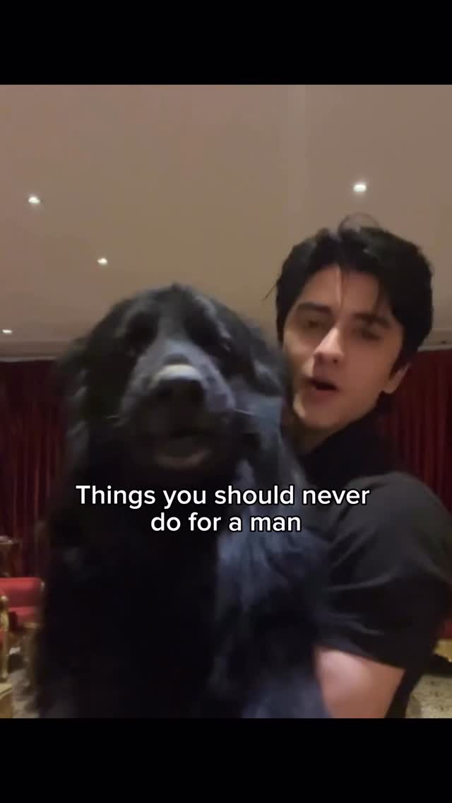 A man dressed in black is holding a large black dog in front of a neutral background lit by soft internal lighting. Overlaid text says, "Things you should never do for a man".