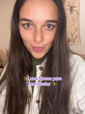 A selfie-style image featuring a woman with long dark hair looking directly at the camera. She is wearing a white button-up top over a green turtleneck. The background is slightly out of focus, with neutral brown and beige tones. Text overlayed on the image states 'Luna Nueva para manifestar' with star emojis.
