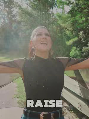 A woman stands outdoors with her arms outstretched, looking upwards with an expression of joy. The background is blurred nature with trees and sunlight. Overlayed text in bold white font at the bottom reads 'RAISE'.