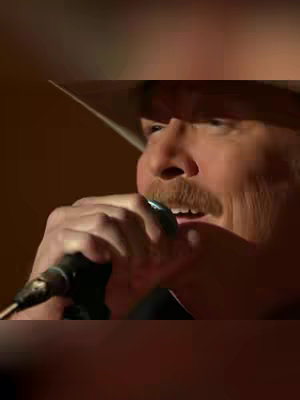 The cover image is a medium shot of a man singing into a microphone. He is wearing a cowboy hat, a dark shirt, and a mustache. The lighting is subdued, suggesting a stage or performance setting. The background is a blurred dark space.