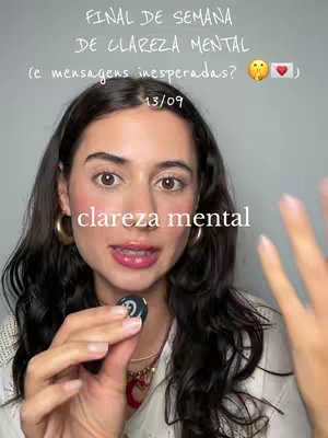 A woman is centered in the frame, with dark curly hair framing her face. She appears to be holding a small object and is mid-sentence. Text overlays appear at the top and bottom of the frame. She has a natural makeup look, with large gold hoop earrings. The background is a neutral, slightly blurred space.
