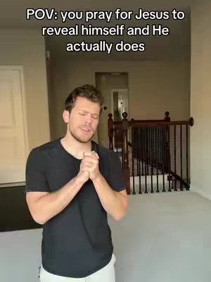 A man in a black t-shirt stands in a house with his hands clasped together in a praying gesture. Overhead text says, 'POV: you pray for Jesus to reveal himself and He actually does'. The background includes a staircase, a door and a glimpse of another room.